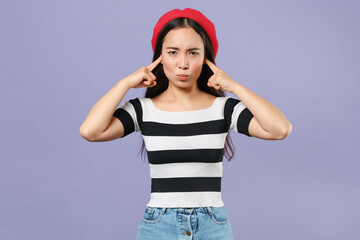 Displeased offended upset young brunette asian woman 20s wearing casual striped t-shirt red beret covering ears with fingers looking camera isolated on pastel violet colour background studio portrait.