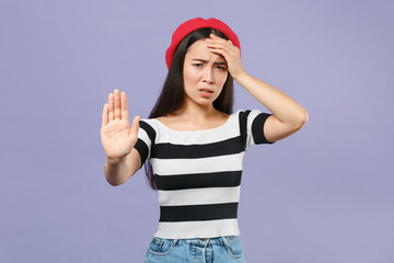 Fototapeta na wymiar Worried dissatisfied young asian woman in striped t-shirt red beret standing showing stop gesture with palm put hand on head looking camera isolated on pastel violet colour background studio portrait.