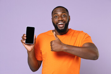Excited young african american man 20s in basic casual orange t-shirt standing showing thumb up hold mobile cell phone with blank empty screen isolated on pastel violet background, studio portrait.