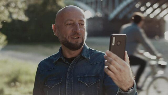 Bald and handsome man with a stylish beard is standing in the park. He has a nice video call with someone he likes. He is very hapy and pleased to have this conversation.