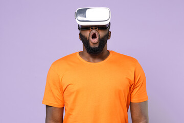 Shocked amazed excited young african american man wearing basic casual orange blank empty t-shirt standing watching in vr headset gadget isolated on pastel violet colour background, studio portrait.