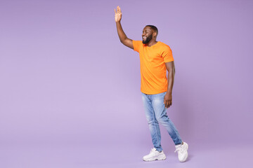 Fototapeta na wymiar Full length side view of joyful young african american man wearing basic casual orange t-shirt waving and greeting with hand as notices someone isolated on pastel violet background, studio portrait.