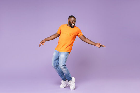 Full length of excited funny young african american man 20s in basic casual orange t-shirt dancing standing on toes pointing index fingers aside isolated on pastel violet background studio portrait.