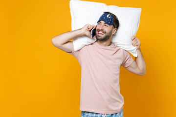 Cheerful young bearded man 20s in pajamas home wear sleep mask hold pillow talking on mobile phone while resting at home isolated on yellow colour background studio portrait. Relax good mood concept.