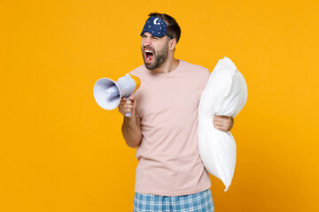 Irritated young bearded man in pajamas home wear sleep mask hold pillow screaming in megaphone...