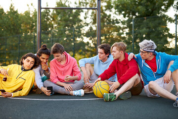 young friends sitting on basketball court, relaxing and taking break after game. talk, laugh, tell...