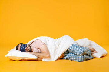 Full length smiling pretty young bearded man in pajamas home wear sleep mask lying with pillow blanket isolated on bright yellow colour background studio portrait. Relax good mood lifestyle concept.