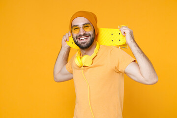 Cheerful smiling young bearded man 20s wearing basic casual t-shirt headphones eyeglasses hat standing hold skateboard looking camera isolated on bright yellow colour background, studio portrait. - Powered by Adobe