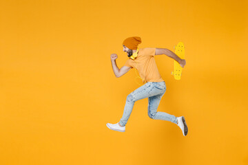 Fototapeta na wymiar Full length side view of cheerful funny young man 20s wearing basic casual t-shirt headphones hat jumping like running hold skateboard isolated on bright yellow colour background, studio portrait.