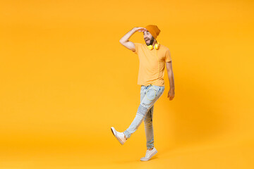 Full length of excited surprised young man wearing basic casual t-shirt headphones hat holding hand at forehead looking far away distance isolated on bright yellow colour background, studio portrait.
