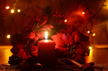 Christmas decorate with red colored candle by placing big red artificial flowers around it and colorful light on background