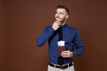 Pensive young traveler tourist business man in blue shirt tie hold passport tickets put hand prop up on chin isolated on brown background studio portrait. Achievement career wealth business concept. © ViDi Studio