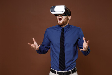 Shocked bearded young business man wearing blue shirt tie watching in vr headset gadget spreading hands isolated on brown colour background studio portrait. Achievement career wealth business concept.