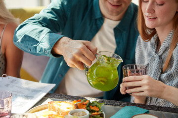 young gallant man pours sweet juice beverage into glasses for women, treats them. in cafe, restaurant