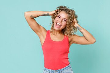 Excited cheerful laughing attractive young blonde woman wearing pink basic casual tank top standing put hands on head looking camera isolated on blue turquoise colour wall background studio portrait.