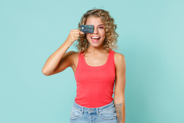 Excited cheerful young blonde woman wearing pink basic casual tank top standing hold covering eye with credit bank card looking camera isolated on blue turquoise colour background studio portrait.