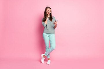 Fototapeta na wymiar Full size photo of interested pretty brown hair woman stand look telephone wear blue jeans sneakers pullover isolated on pink background