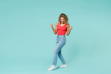 Full length side view of joyful young blonde woman 20s in pink tank top jeans standing clenching fists doing winner gesture keeping eyes closed isolated on blue turquoise background, studio portrait.