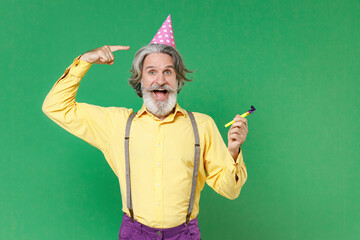 Excited cheerful elderly gray-haired mustache bearded man in yellow shirt suspenders pointing index finger on birthday hat hold pipe looking camera isolated on green colour background studio portrait.