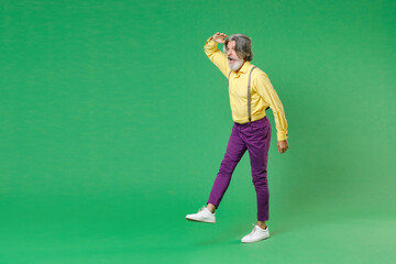 Full length side view of shocked elderly gray-haired mustache bearded man in yellow shirt suspenders holding hand at forehead looking far away distance isolated on green background, studio portrait.