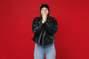 Shocked amazed young brunette woman 20s wearing basic casual black leather jacket white t-shirt hat covering mouth with hands looking camera isolated on bright red colour background studio portrait.