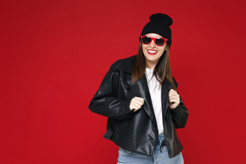 Smiling cheerful beautiful young brunette woman 20s wearing basic casual black leather jacket white t-shirt hat dark eyeglasses looking camera isolated on bright red colour background studio portrait.