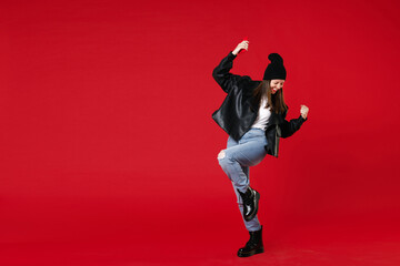 Full length of happy young brunette woman 20s wearing black leather jacket white t-shirt hat using mobile cell phone doing winner gesture clenching fists isolated on red background studio portrait.