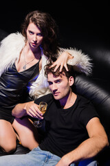 Seductive brunette woman holding hand on boyfriend head, while sitting on knees on sofa in nightclub