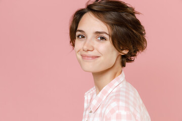 Side view of smiling pretty cheerful charming cute young brunette woman 20s wearing casual basic checkered shirt standing looking camera isolated on pastel pink colour background, studio portrait.