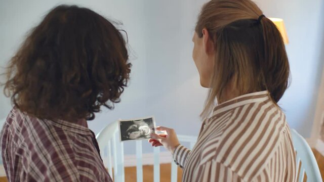 Back view of young married gay couple holding sonogram picture standing in nursery room with crib