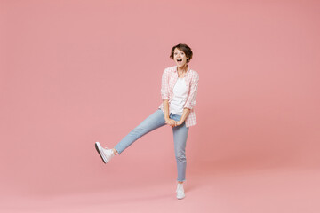 Fototapeta na wymiar Full length of excited cheerful funny young brunette woman 20s wearing casual checkered shirt standing dancing rising leg keeping mouth open isolated on pastel pink colour background, studio portrait.