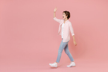 Fototapeta na wymiar Full length side view of excited cheerful young woman 20s wearing casual basic checkered shirt waving greeting with hand as notices someone isolated on pastel pink colour background, studio portrait.