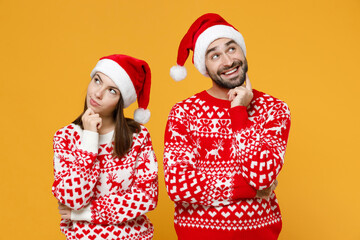 Pensive young Santa couple friends man woman 20s in red sweater Christmas hat put hand prop up on chin isolated on yellow background studio portrait. Happy New Year celebration merry holiday concept.