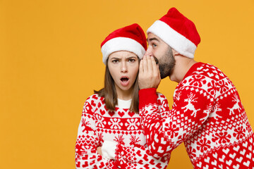 Shocked young Santa couple friends man woman in red sweater Christmas hat whispering secret behind...