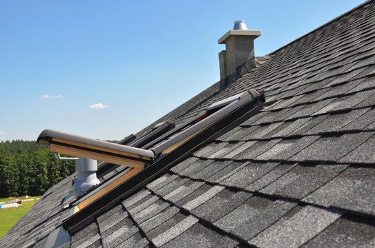 A gray roof covered with dimensional architectural asphalt shingles with an attic skylight, ventilation pipe, air duct and chimney. A close-up on an open attic skylight.