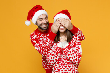 Cheerful funny young Santa couple friends man woman 20s in red sweater Christmas hat covering eyes with hands isolated on yellow background studio. Happy New Year celebration merry holiday concept.