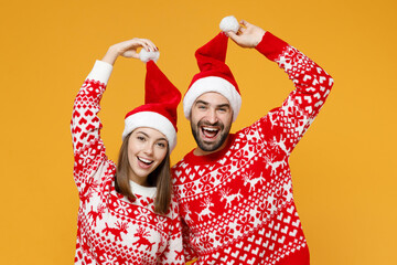 Cheerful funny laughing young Santa couple friends man woman 20s wearing red sweater hold Christmas hat isolated on yellow background studio portrait. Happy New Year celebration merry holiday concept.