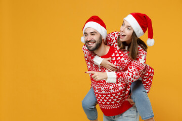 Excited young Santa couple friends man woman in Christmas hat giving piggyback ride to joyful, sit...