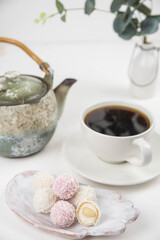 Fototapeta premium White coconut candy balls on handmade plate. Coconut cookies on white background with the cup of tea. Teapot with black tea. 