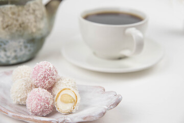 White coconut candy balls on handmade plate. Coconut cookies on white background with the cup of tea. Teapot with black tea. 