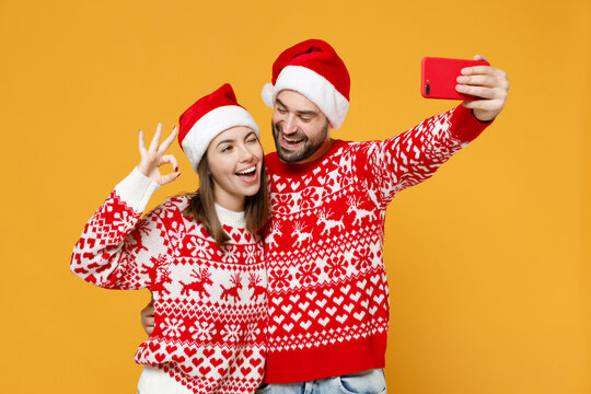 Cheerful young Santa couple friends man woman in sweater, Christmas hat doing selfie shot on mobile phone showing Ok gesture isolated on yellow background. Happy New Year celebration holiday concept.