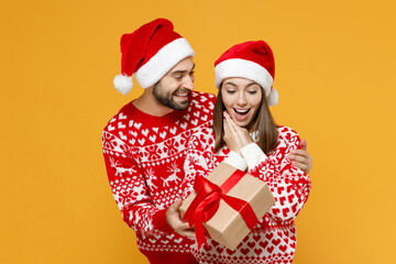 Shocked young Santa couple friends man woman 20s in red sweater Christmas hat hold present box with gift ribbon bow isolated on yellow background. Happy New Year celebration merry holiday concept.