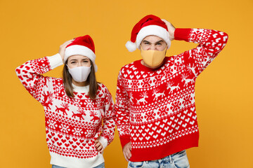 Worried Santa couple friends man woman in sweater Christmas hat face mask to safe from coronavirus covid-19 put hands on head isolated on yellow background. Happy New Year celebration holiday concept.