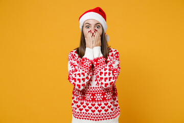 Shocked young Santa woman 20s in red sweater Christmas hat covering mouth with hands looking camera isolated on yellow background, studio portrait. Happy New Year celebration merry holiday concept.