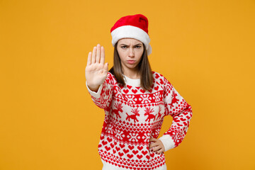 Dissatisfied young brunette Santa woman 20s in red sweater Christmas hat showing stop gesture with palm isolated on yellow background studio portrait. Happy New Year celebration merry holiday concept.