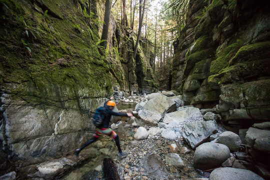 Man exploring mossy dark canyon leaps across river.