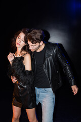 Man in leather jacket embracing sexy girlfriend in dress on black