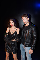 Sexy brunette woman with crossed arms standing near man in leather jacket with backlit on black