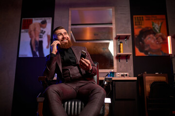 young male in suit, barber man hold trimmer in hands and talk on phone, at work place. in dark room