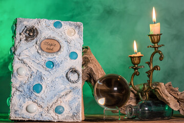White magic book, crystal ball and burning candle on the table on the green smoke background.
